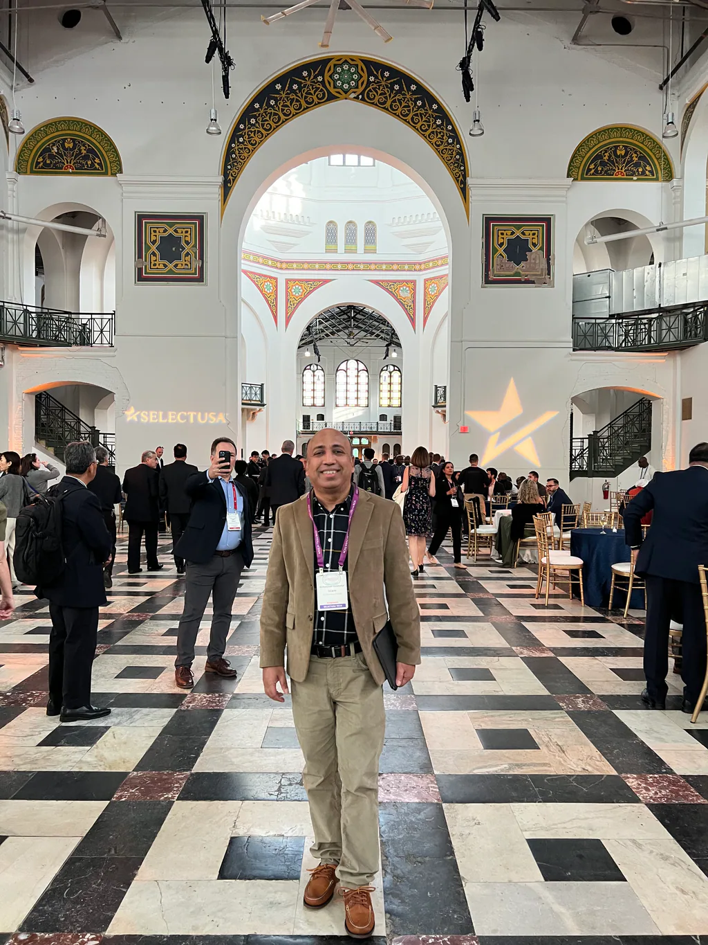 Full-length portrait in the marble hall of a professional event venue, delegates in the background.