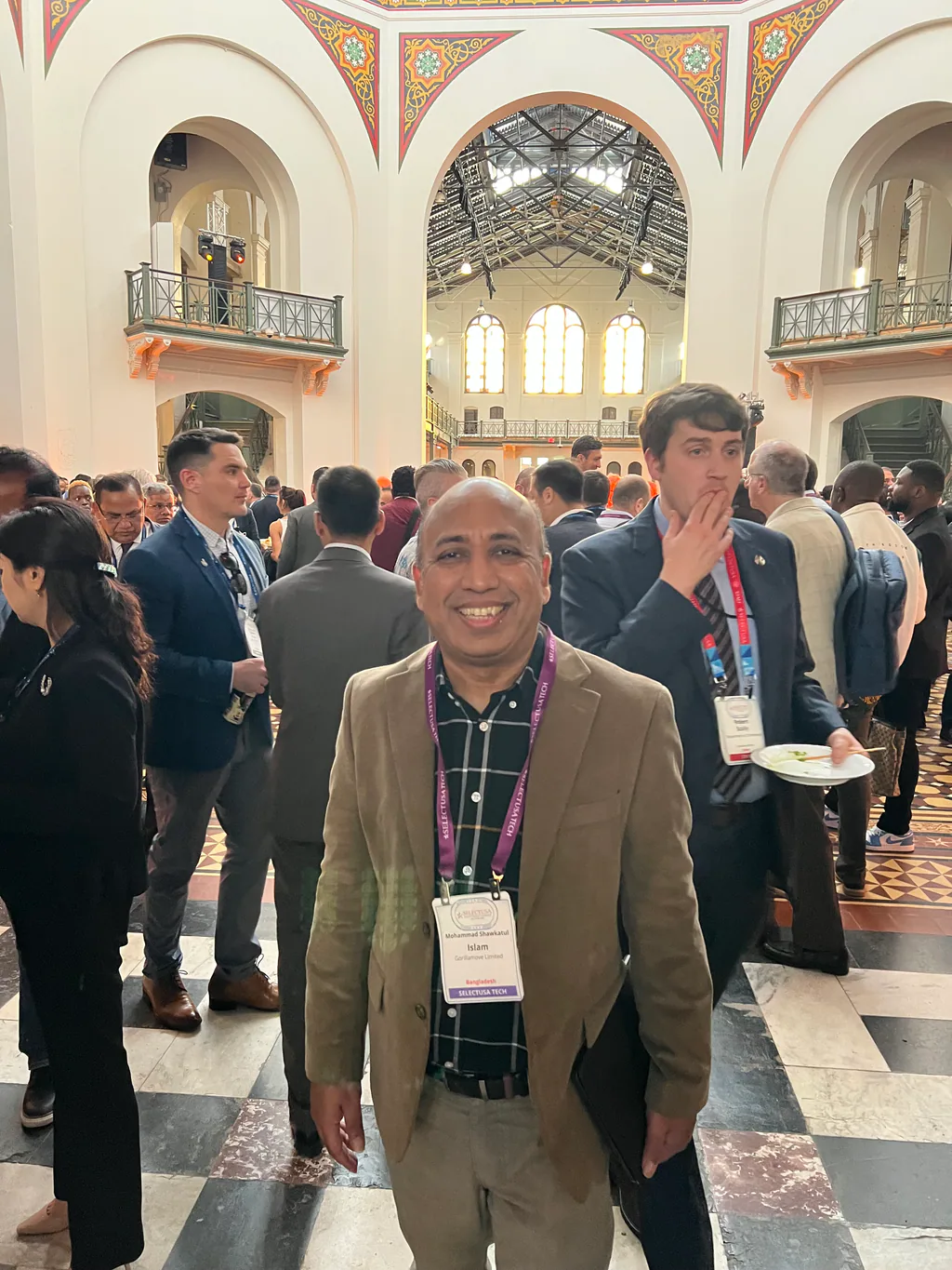 Close portrait at a conference reception, delegates and a historic hall interior behind.