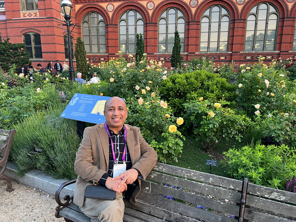 Seated portrait on a bench in a rose garden beside a historic brick building.