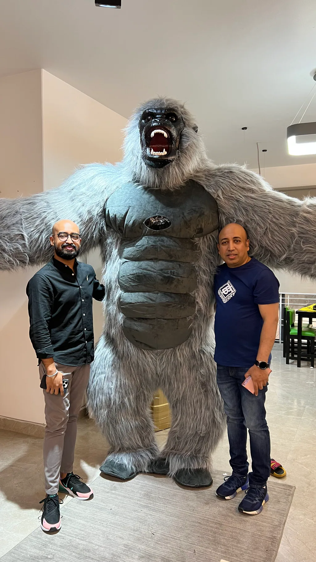Shawkat Islam and Sadman Islam standing either side of a large Gorilla mascot with extended arms, in an office atrium.